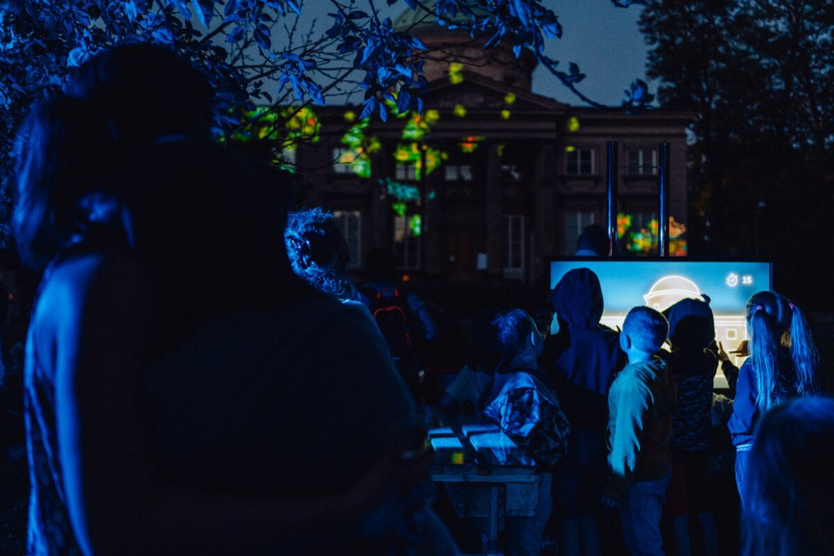 In the evening, a group of people, including children, gather outside for a photo report of the event. They focus on a brightly lit screen displaying colorful images. A building is visible in the background, and trees partially obstruct the view. Dim lighting and shadows create a nocturnal atmosphere.   