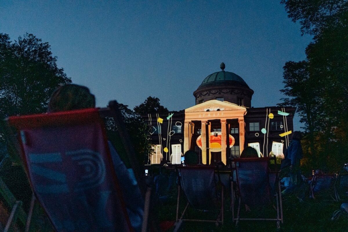 A few people relax on deck chairs at dusk in front of a large building with a dome, columns and illuminated abstract projections on the facade. Surrounding trees encircle the scene, and the sky is a dark blue, suggesting night has fallen; it's a perfect photo-op of the event. 