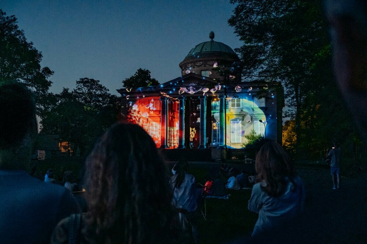 People gather in the evening for a photo event to watch the projection of vibrant light on the façade of a historic building with a domed roof. The projections display various colorful patterns and images, illuminating the structure against the darkening sky. 