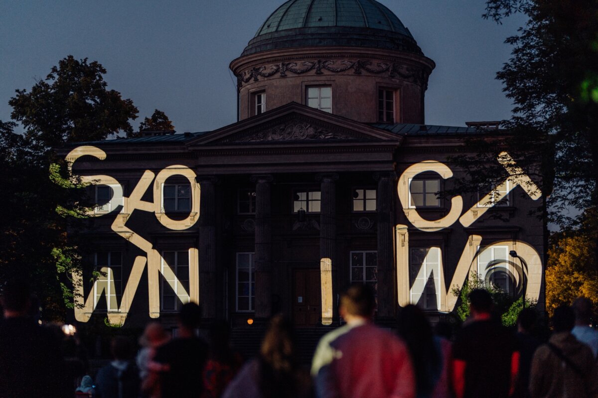 A night stage in a historic building with a domed roof and columns. It has an illuminated projection with the words "SLOW" and "C O W N". The foreground shows silhouettes of people watching the exhibition. Trees surround the building on both sides, creating a captivating photo-op of the event.   