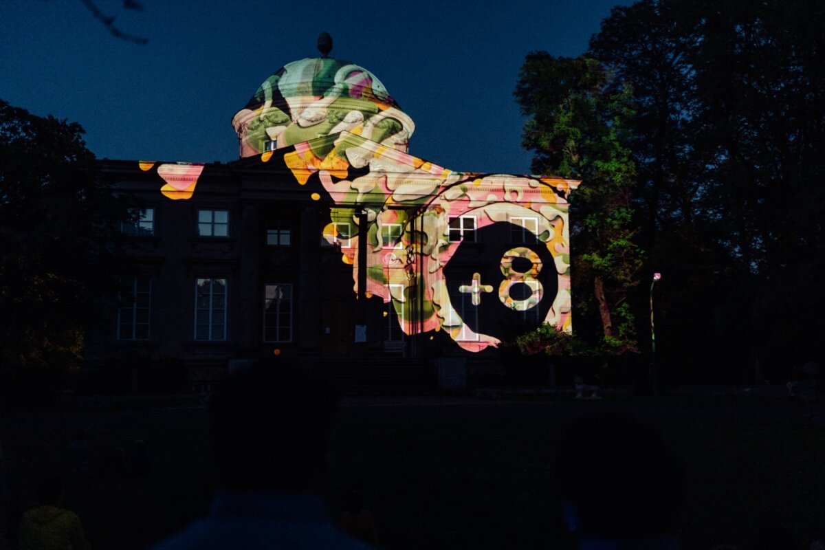 The building, with its classical architecture, is illuminated at night with colorful light projections. Photo coverage of the event shows a variety of abstract designs, including geometric shapes and vivid patterns, casting a captivating glow against the dark trees and sky. 