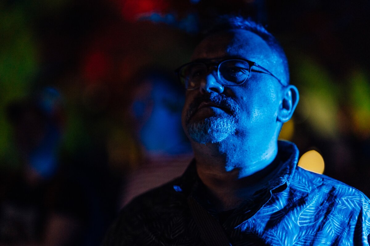 A man wearing glasses and a short beard is standing in a dimly lit, colorful environment, illuminated mainly by blue and dim yellow light. He appears to be looking slightly upward and is wearing a patterned shirt. The blurred background features a variety of hues, creating an atmospheric snapshot of the event.  