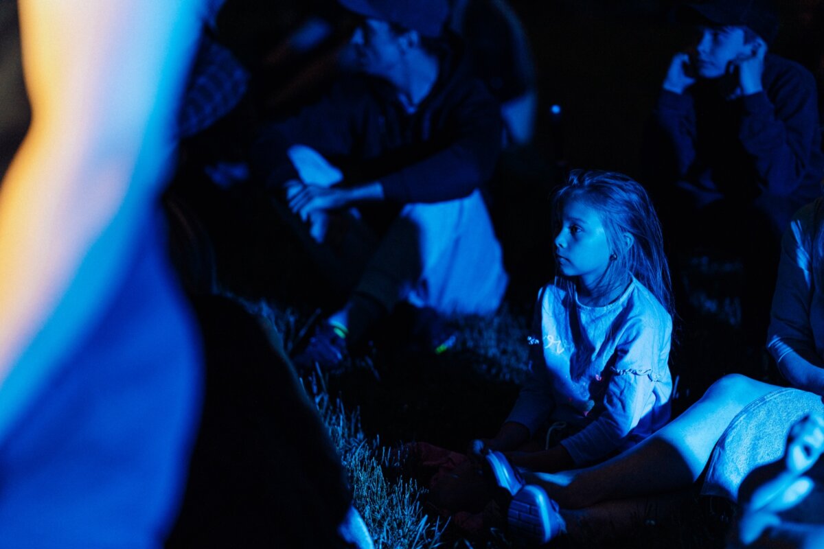 A young girl, illuminated by a blue light, sits on the grass among other people during what appears to be a nighttime outdoor event. She looks intently to the left of the stage, while others around her seem equally focused - shots from a photo essay of the event showing the crowd involved. 