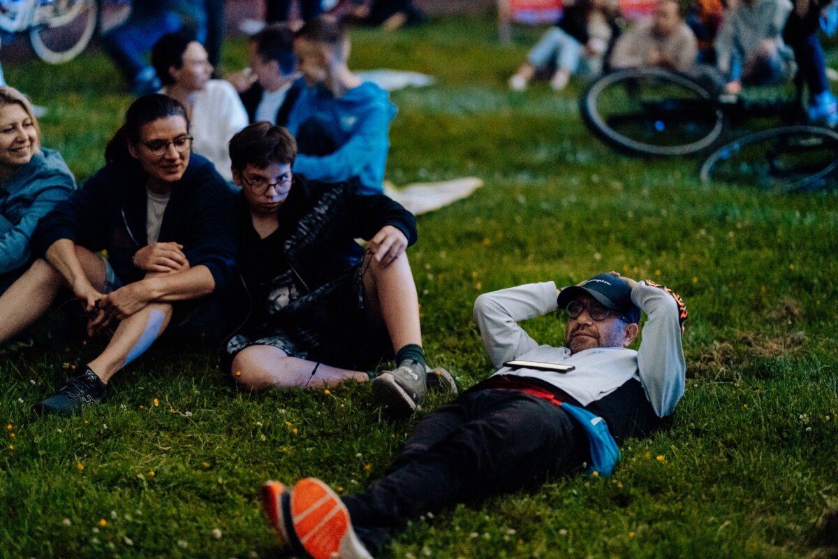 A group of people enjoying a casual outdoor party. Two people are sitting on the grass, smiling and looking toward something off-camera. Next to them on the grass lies a person wearing a hat with his hands folded behind his head, relaxed and content. In this photo report of the event, bicycles can be seen in the background.   