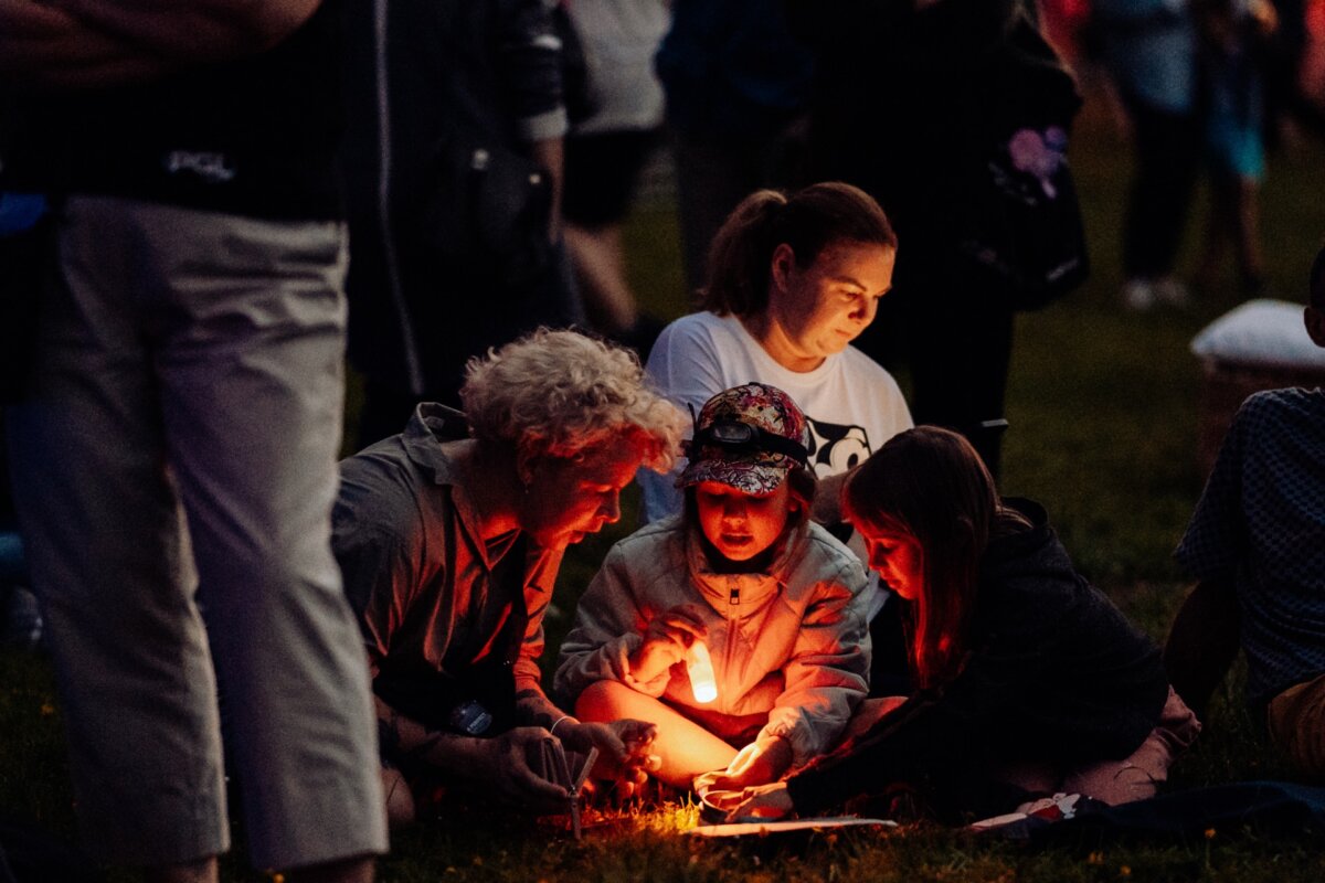 At dusk, a group of children and an adult are sitting on the ground, illuminated by a small lamp they are holding. In the background, the scene is full of people and it seems that everyone is engaged in some outdoor activity or event, capturing a perfect photo-reportage of the event. 