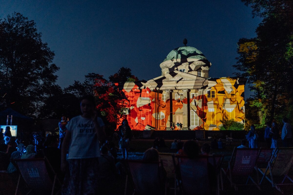 Pictured is a nighttime outdoor event with the historic building illuminated. The facade of the building is lit up with colorful abstract projections. People gather in front of the building, sitting on chairs and on the ground, enjoying the vibrant display - a perfect photo-op of the event.  