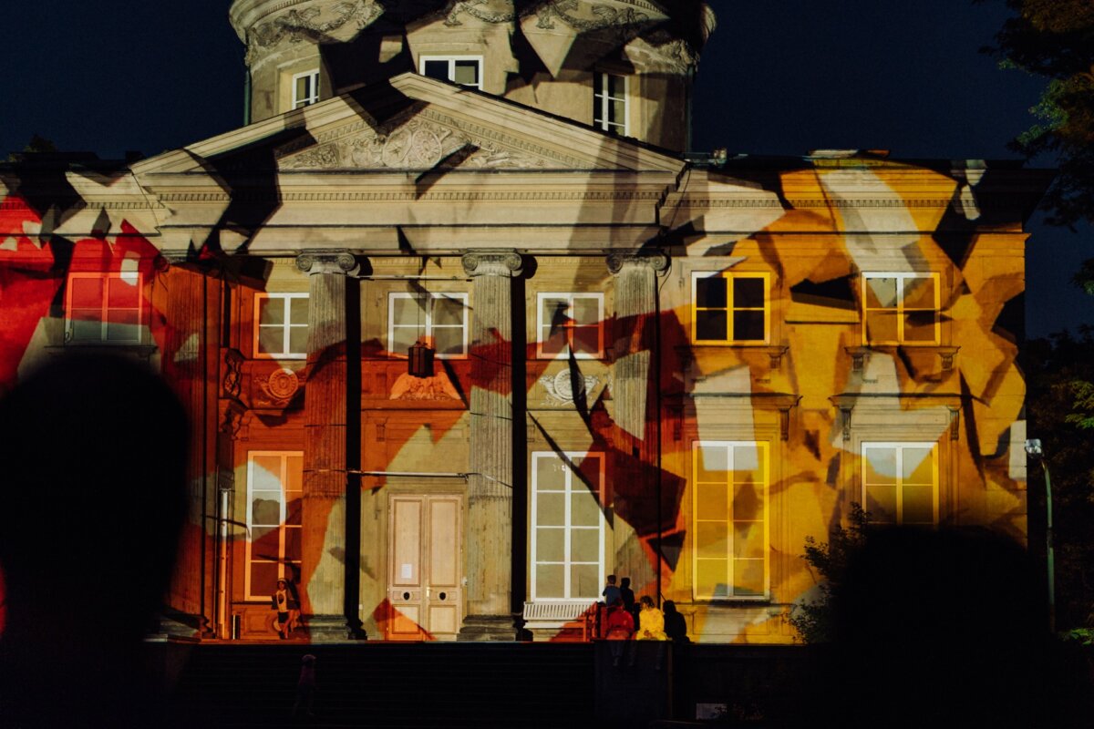 The grand historic building is illuminated at night with colorful abstract light projections. The patterns in shades of red, orange and yellow create a striking effect on the facade. In the foreground you can see silhouettes of people observing the exhibition - a perfect photo-op of the event.  