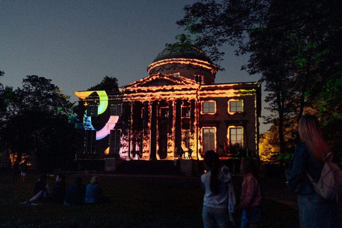 The historic building is illuminated at night with colorful, abstract light projections. Images on the facade include vivid swirls and geometric shapes. Photo coverage of the event shows several people in the foreground, some sitting, some standing, observing the exhibition.  