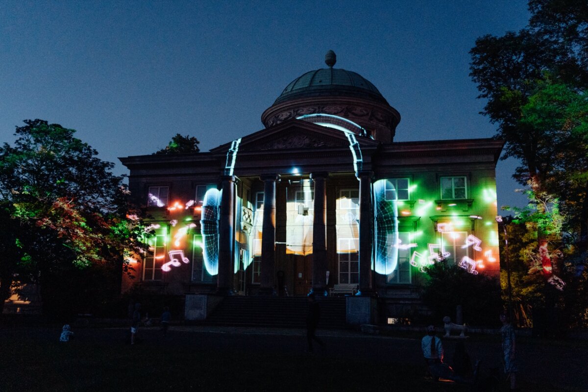 A stately building with a domed roof is illuminated by colorful projections of notes and speakers against a sunset sky. The building is surrounded by trees, and people are scattered on the lawn watching the light show, creating the perfect stage for photo coverage of the event. 