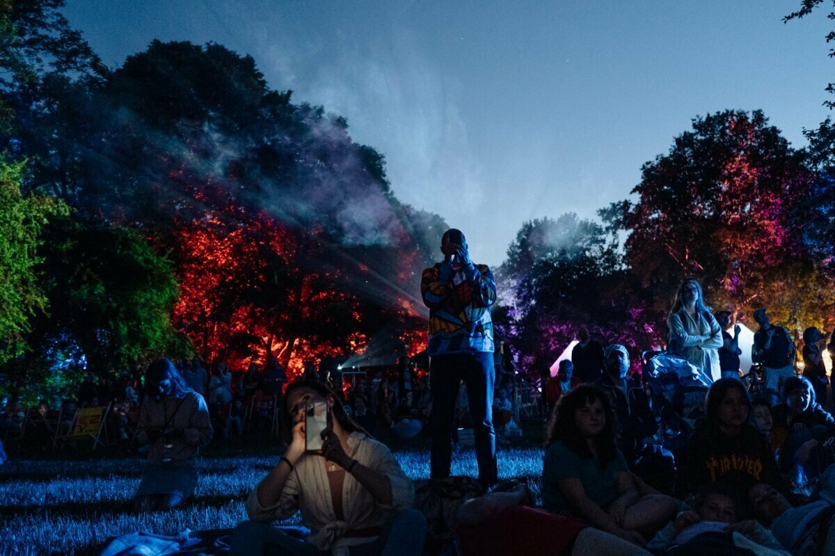 People gather outside at night, sitting and standing on the lawn surrounded by trees illuminated by colored lights. The sky is darkening, suggesting dusk or night. Some are taking photos for a photo essay of the event, while others are simply enjoying the atmosphere.  