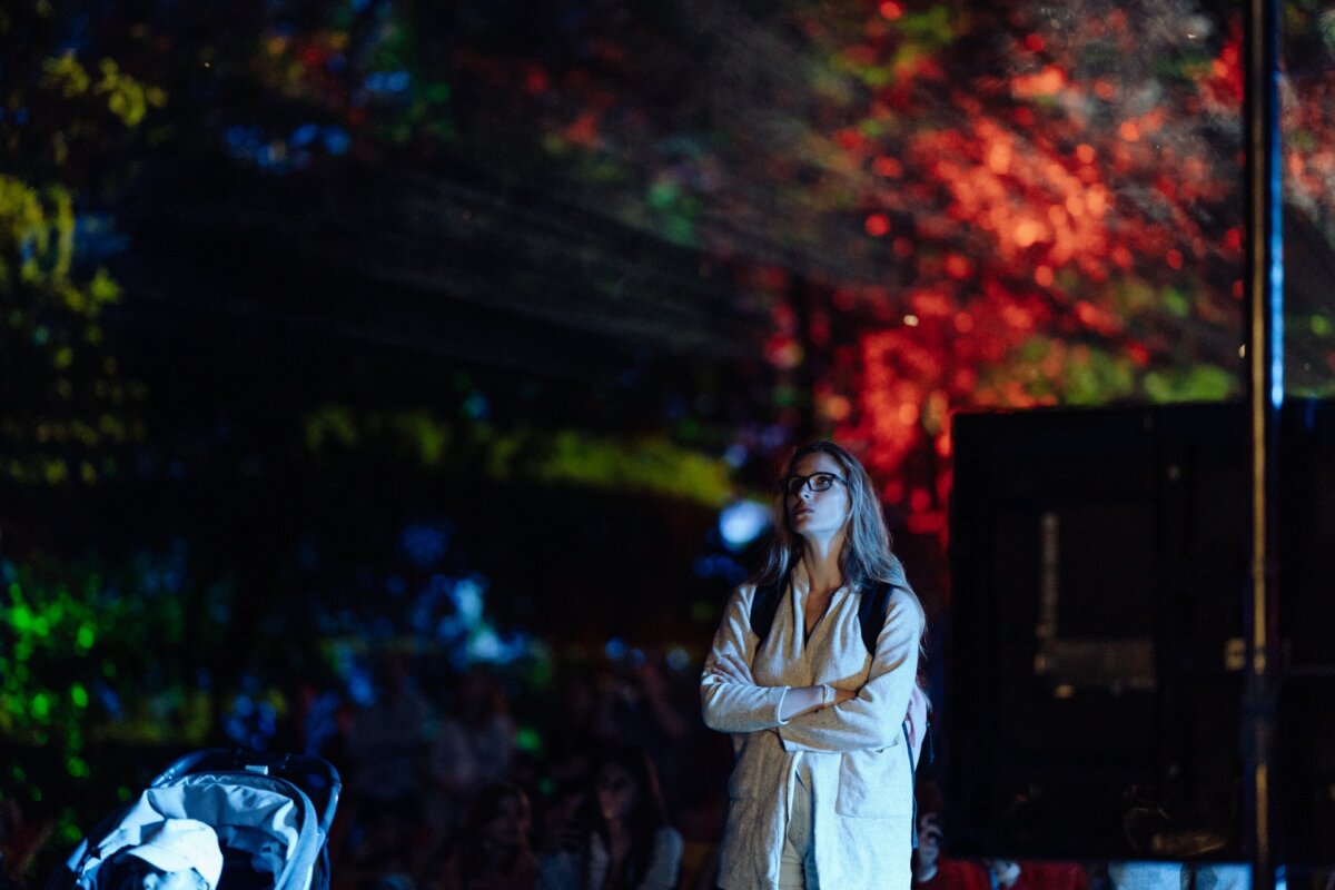 A person with long hair and glasses stands with his arms crossed and looks at a mesmerizing light show in a dark outdoor environment. The background is illuminated with colored lights, creating an abstract pattern. On the left, a cart is visible - perfect for photo coverage of the event.  