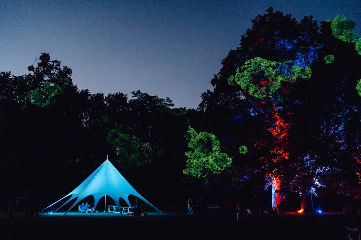 A night scene showing a large white tent illuminated by blue lights, set in a wooded area. The trees around the tent are decorated with colorful lights in shades of blue, red and green, creating a lively atmosphere against the darkening sky. The perfect setting for a photo event.  