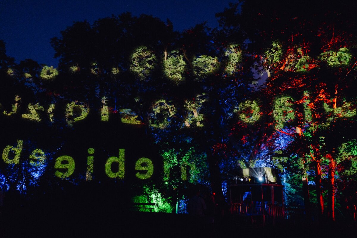 A night scene of trees illuminated by colored lights, including green and red. The displayed text is visible on the trees, creating an artistic pattern. In the background is a small structure with lights, partially obscured by dense vegetation - a spectacular photo-recording of the event.  