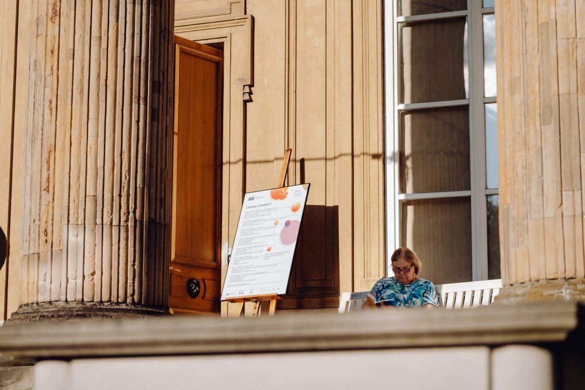 A person with glasses sits on a bench and reads next to a large signboard on an easel. In this event photo essay, the scene is set in front of a large building with tall columns and open wooden doors. Sunlight casts shadows, and a multi-paned window can be seen in the background.  