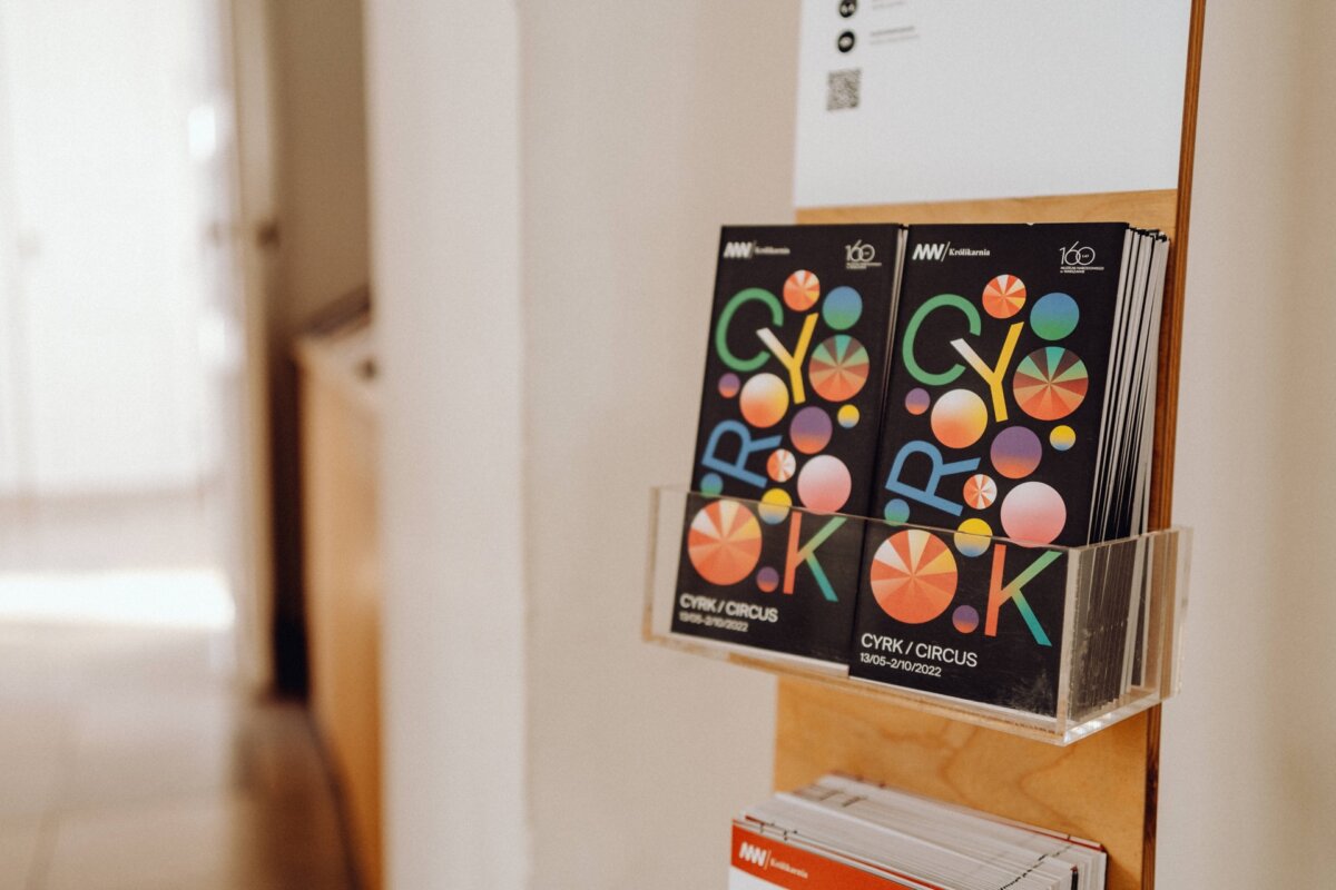 The brochure rack contains multiple copies of a magazine titled "CIRK/CIRCUS" in bold letters, with a colorful geometric circular pattern on the cover. Situated in a well-lit area next to a wooden surface, it offers an inviting display of photo stories from the event. 