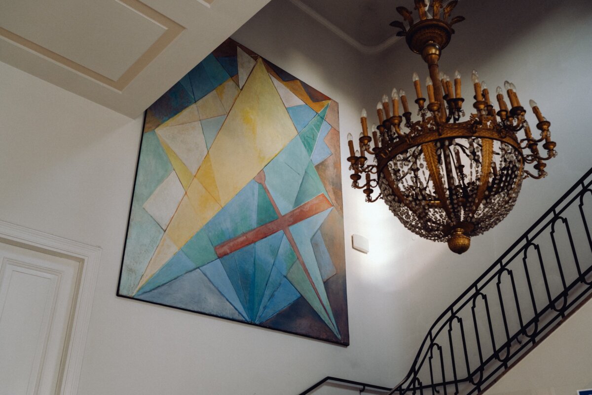 A large chandelier with intricate details hangs on the well-lit staircase. On the wall next to the staircase, an abstract geometric painting of various colorful shapes is displayed, which will add a modern, artistic touch to a classic interior, perfectly suited for a photo-op of the event. 