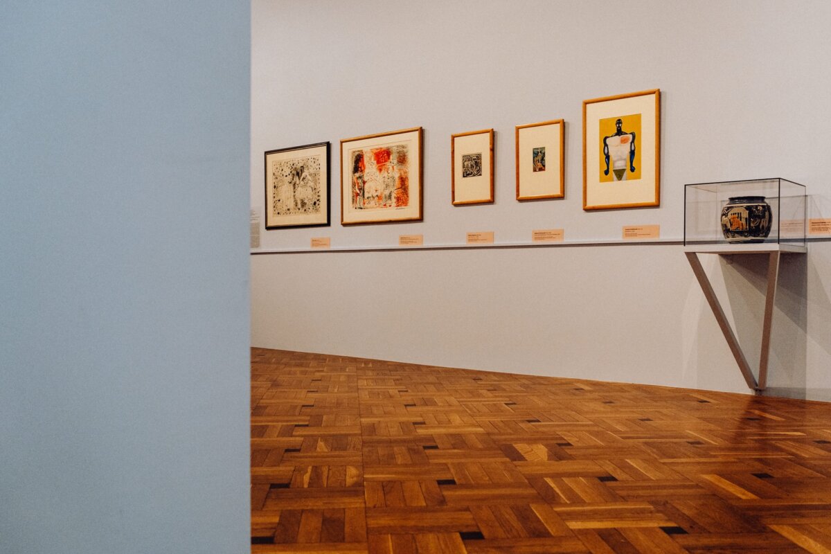 A gallery room with a floor lined with wooden parquet, where a series of framed artworks are displayed on a white wall. The artworks vary in size and style, alluding to the event's photo-op. A glass display case sits on a shelf to the right. The room is empty and well lit.   