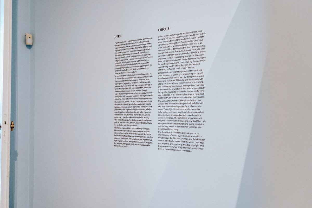 A white wall with text in two columns, titled "CIRK" and "CIRKUS," describes the history, aspects or definition of circuses. The exhibit is part of the display, with the doorframe on the left. The well-lit wall gives the impression of being part of a photo essay of the event.  