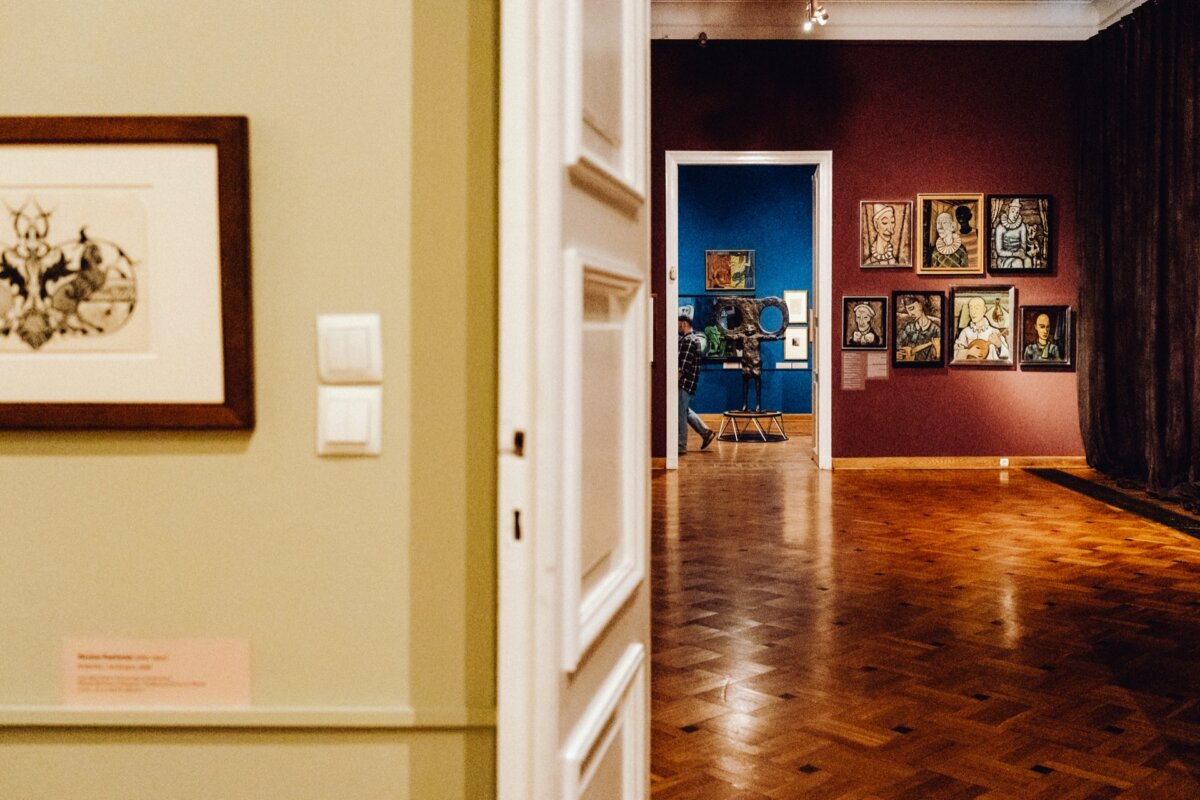 A view of the art gallery through an open doorway, displaying framed paintings on the walls in vibrant colors. The corridor is warmly lit, revealing a wooden floor and a variety of artwork displayed on walls painted in shades of green, blue and burgundy - a perfect photo-op of the event. 