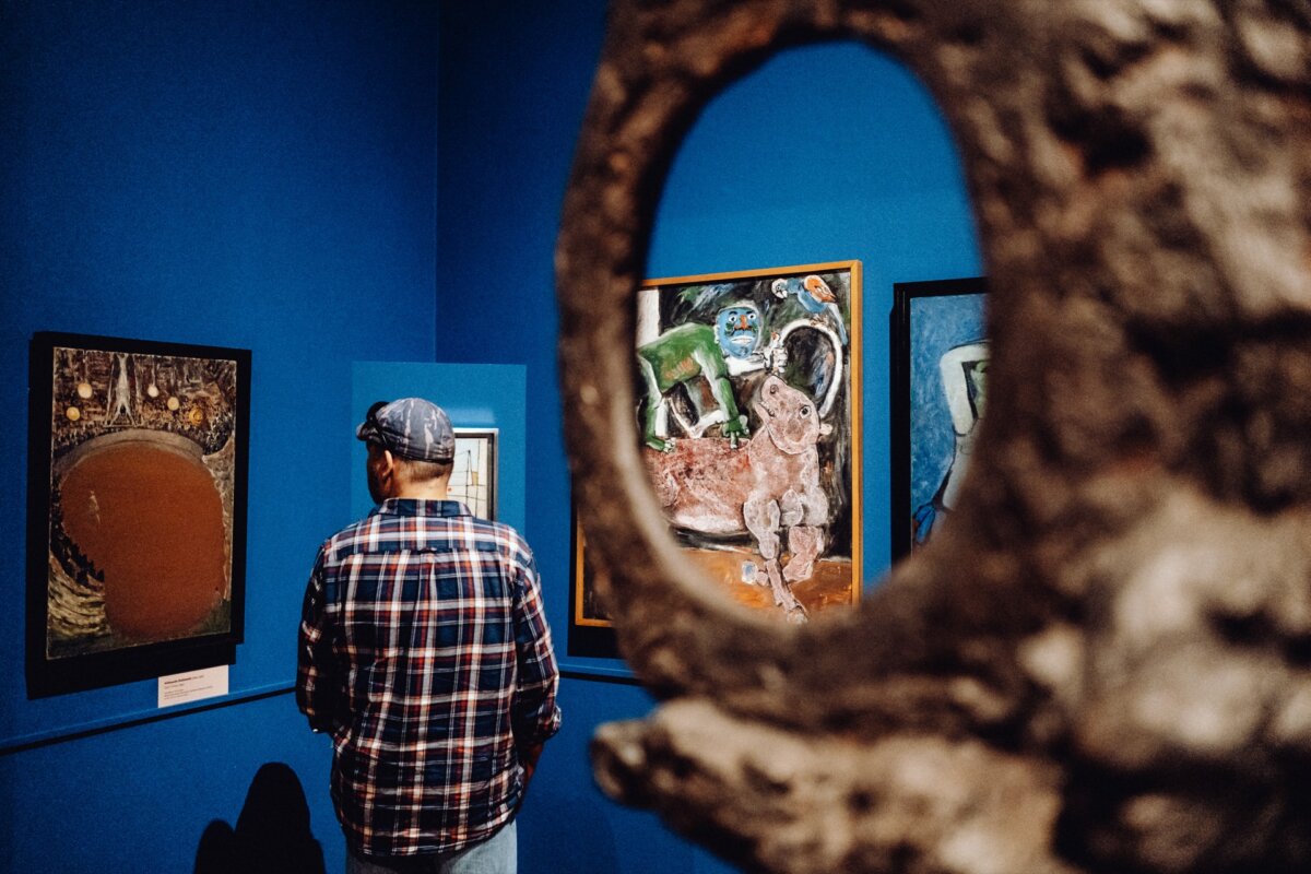 A person dressed in a checkered shirt and cap stands in an art gallery with blue walls, engrossed in abstract paintings. Part of a large sculpture with a circular opening surrounds the person and part of the artwork, creating an intriguing Photo Event. 