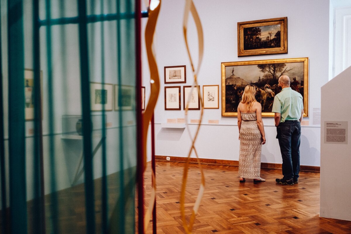 A man and a woman observe framed works of art on the walls of a museum gallery with wooden floors and light blue walls. They give the impression of being deeply involved in viewing or discussing the paintings and drawings, creating an atmosphere of almost photo-reportage of the event. 