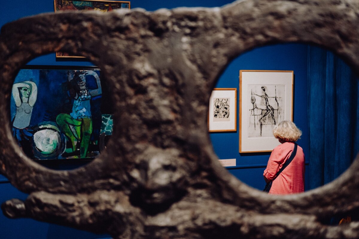 A person with light hair, wearing a pink shirt, stands in an art gallery and looks at various paintings and drawings on a blue wall. In this Event Photo Report, a fuzzy sculpture with large holes through which the person can be seen can be seen in the foreground. 