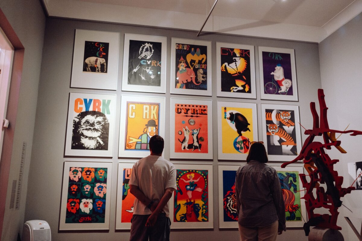 Two people stand in a gallery and observe a collection of colorful retro-style posters depicting various animals and circus themes. The posters are arranged in a grid on a white wall. To the right of the people, a modern art sculpture can be seen, capturing the essence of the event's photo-op.  