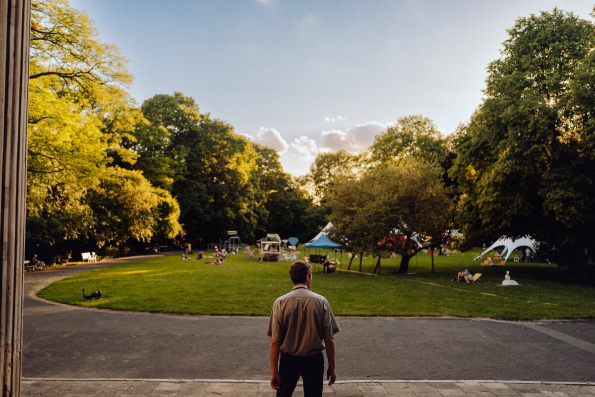 A person stands on a sidewalk and observes a peaceful scene in a park. Trees with lush green leaves surround a grassy area where people are relaxing and enjoying being outdoors. Tents are set up in the background, suggesting a photo-op of an event, or casual gathering.  