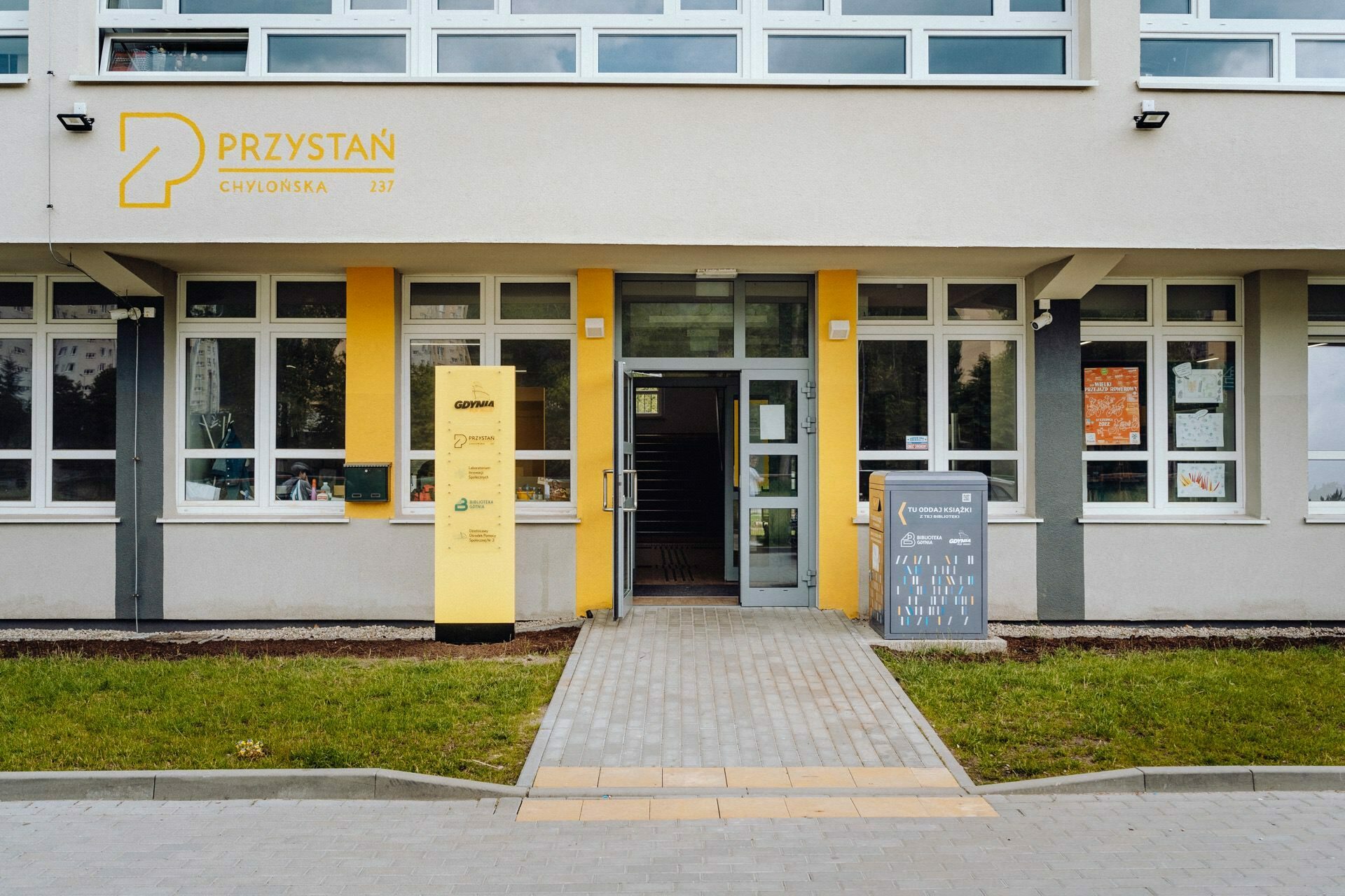 The modern entrance to the building, with its yellow door and matching yellow columns on the gray facade, is perfect for a Warsaw-based photographer to capture. Mailboxes on the right side, green grass in front and large windows on the upper floors complement the "PRZYSTAŃ" sign exposed in yellow lettering above the door. 