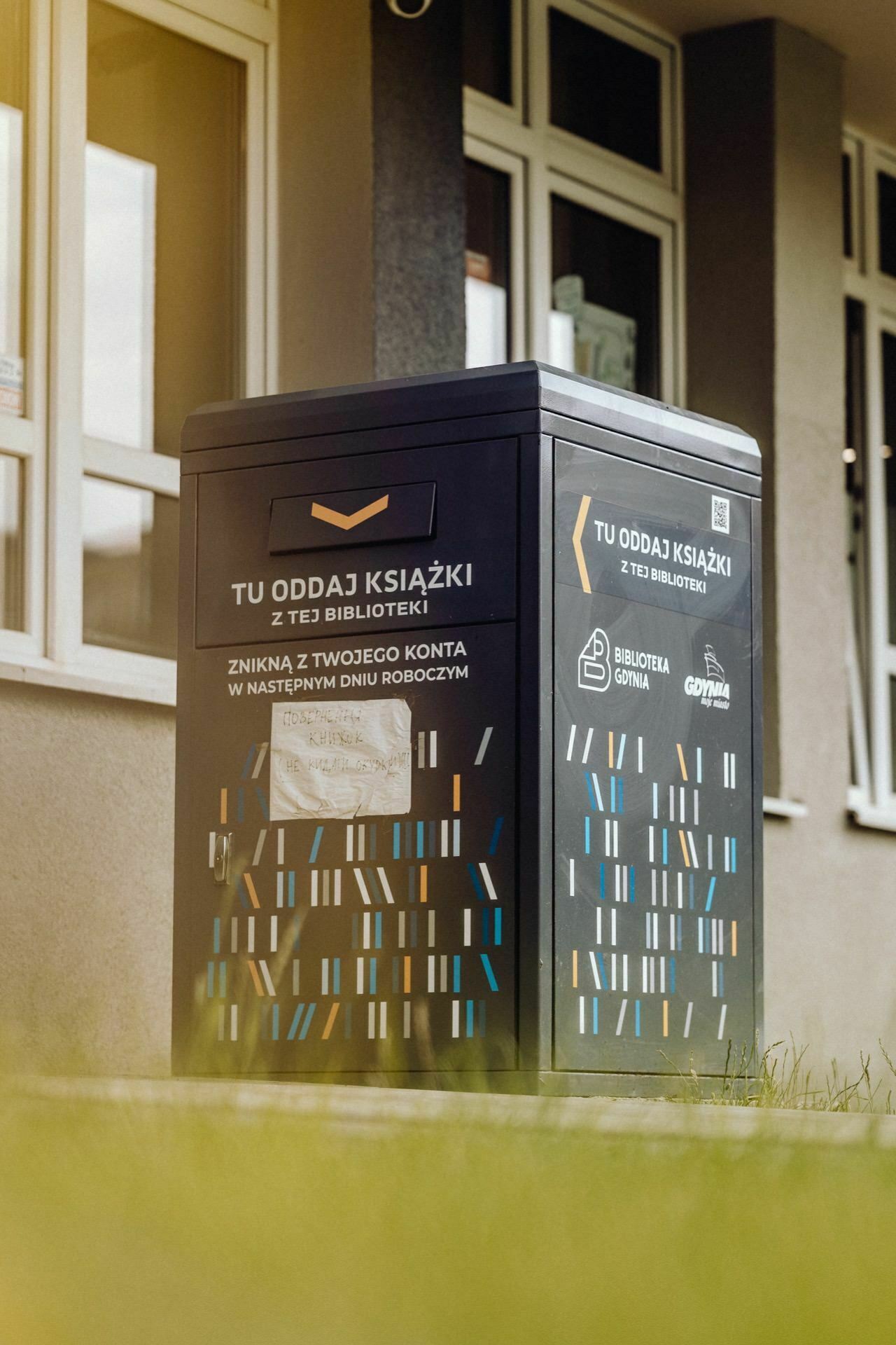 A dark blue book return box, captured by a photographer from Warsaw, stands outside a building with several windows. The box contains text in Polish stating that it is for returning books to the library. The ground around the box is overgrown with yellowish-green grass.  