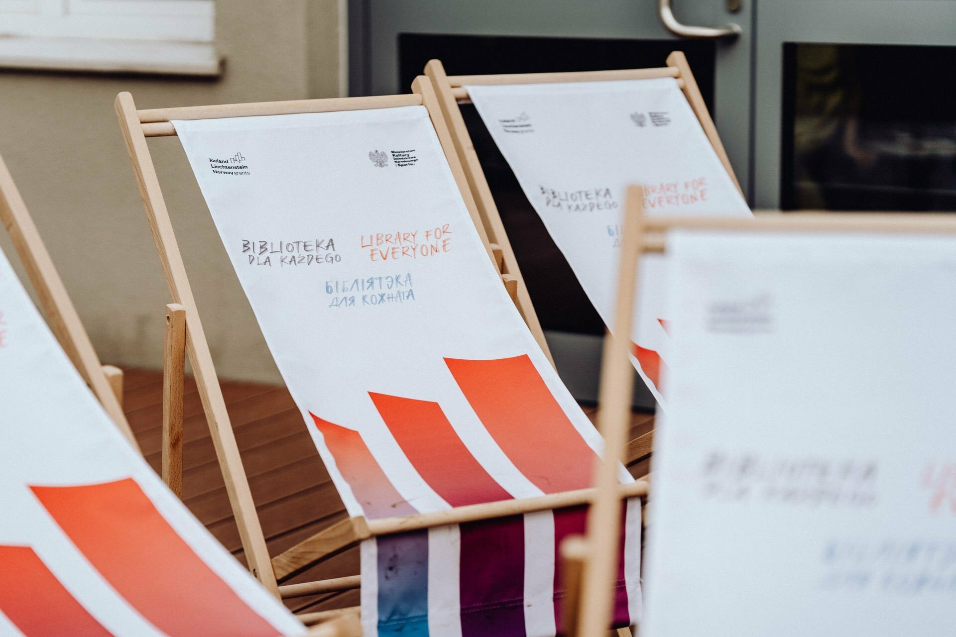 A set of white fabric recliners with wooden frames arranged on a wooden floor. The chairs feature multilingual text, including "Library for Everyone" and "Бібліотека для кожного," as well as colorful abstract patterns in red and orange. Perfect for your next photo event.  