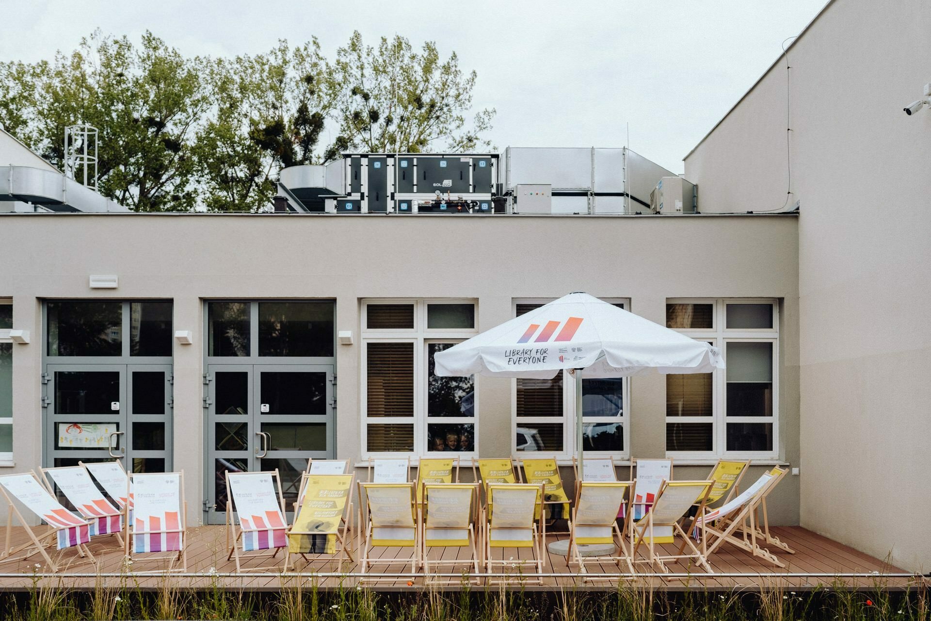 The outdoor patio has several yellow deck chairs and small tables, under a white umbrella with red and orange stripes. The patio, perfect for any Warsaw photographer to capture moments, is adjacent to a building with large windows and doors, with greenery visible in the background. 