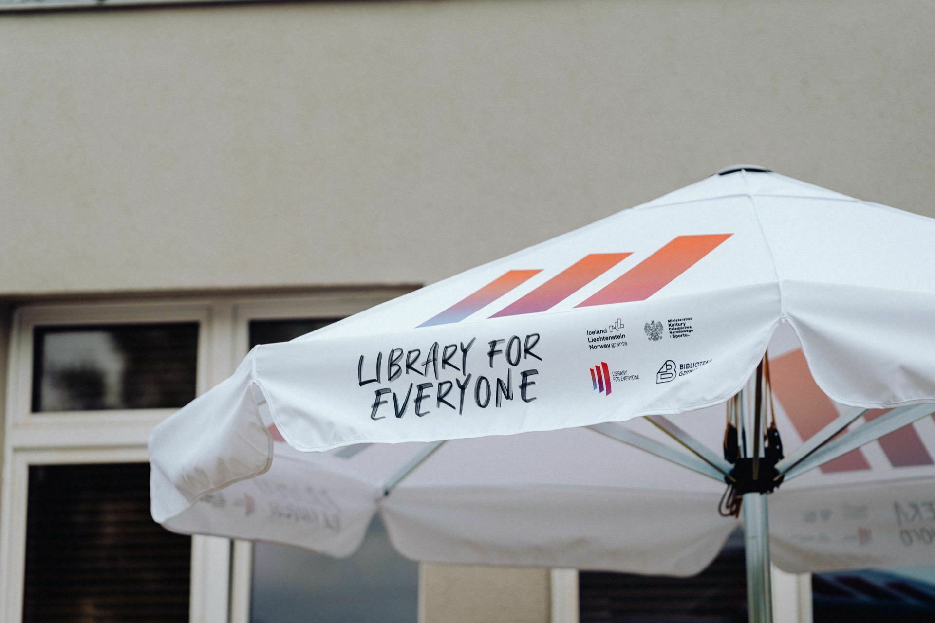 White garden umbrella with the words "Library for everyone" printed on the side with black text, logos and symbols. The umbrella with colorful stripes, placed in front of the building with windows, is perfect for capturing in a photo report of the event. 