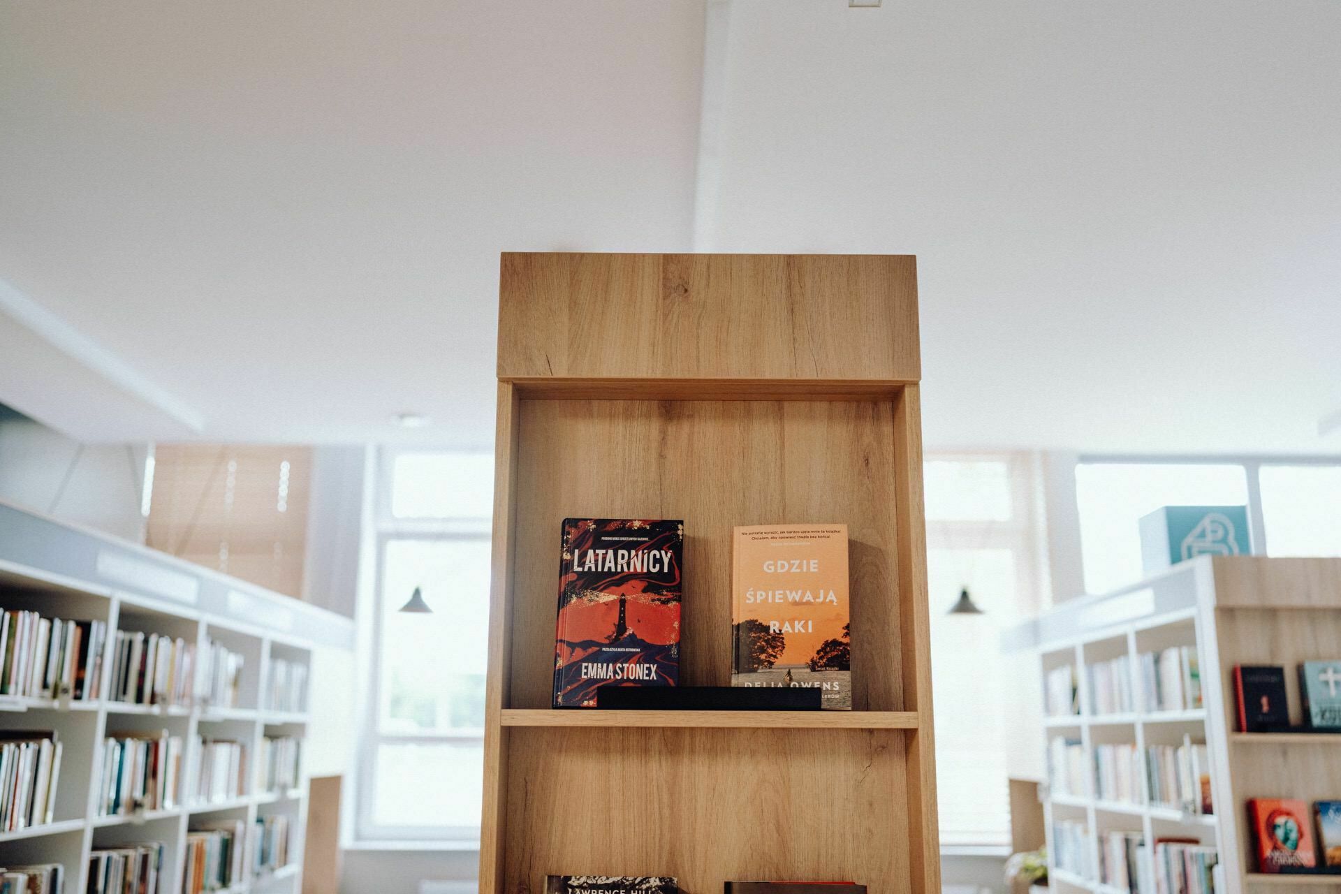 A wooden bookcase in a well-lit library displays two books, as if captured by a photographer from Warsaw. The book on the left has a dark cover with light text that reads "Lighthouse Keepers" by Emi Stupchev. On the right is "Where the Crayfish Sing," in a lighter cover with an illustration showing a sunset.  