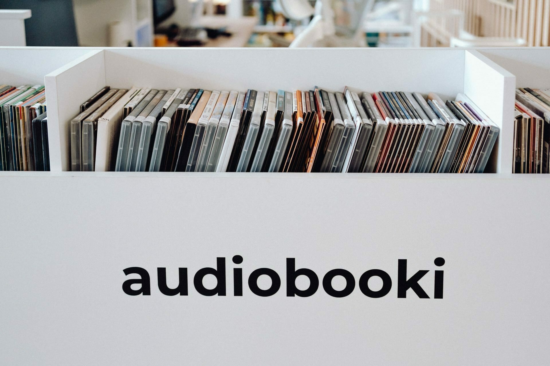 A bookshelf labeled "audiobooks" contains a variety of audiobooks in CD format. The carefully organized collection features spines in a variety of colors and styles, indicating a diverse selection of titles. In the background, a softly blurred photo of the event complements the decor of the library or store.  
