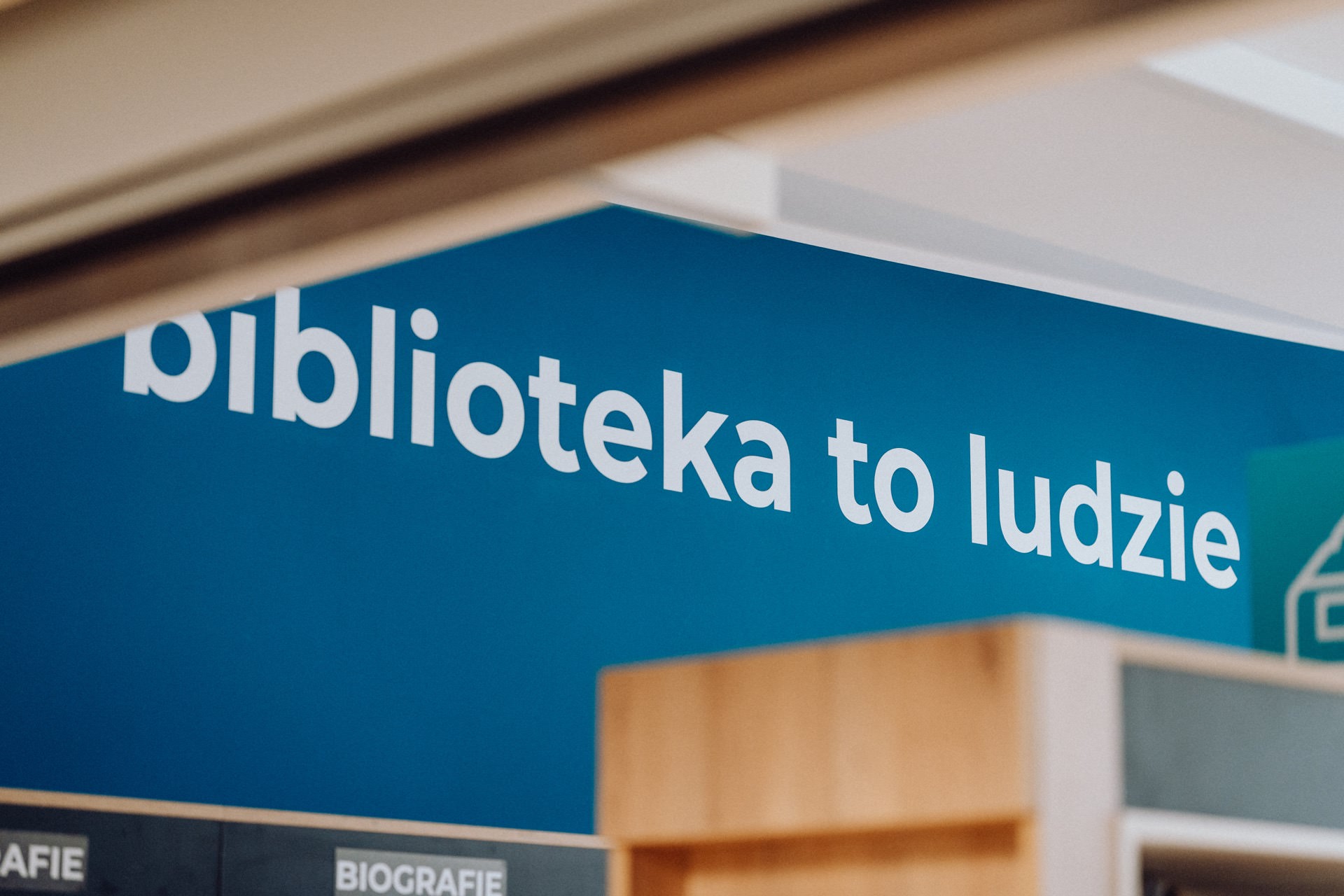 On the blue wall of the library is the white text "library for the people," which in Polish means "library is people." Below the text, a section of a wooden bookshelf and a section titled "BIOGRAPHIES" are visible, perfectly captured by a photographer from Warsaw. 