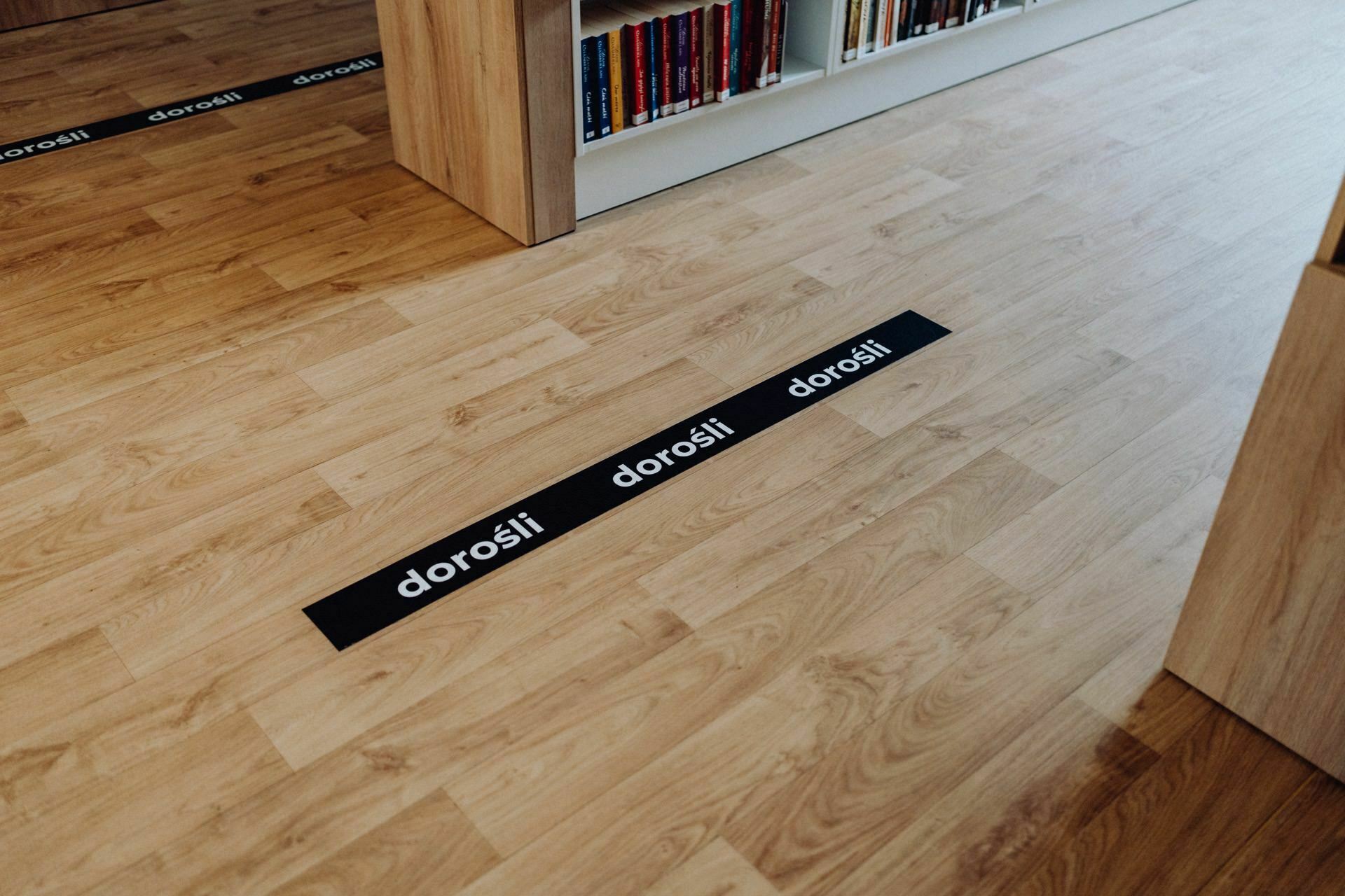 Wooden floor of the room with a bookshelf visible in the background. Black and white text "adults" appears on the floor stickers, arranged in a line leading toward the bookshelf. The scene, captured by a Warsaw-based photographer, depicts a path or guideline, likely to be found in a library or bookstore.  