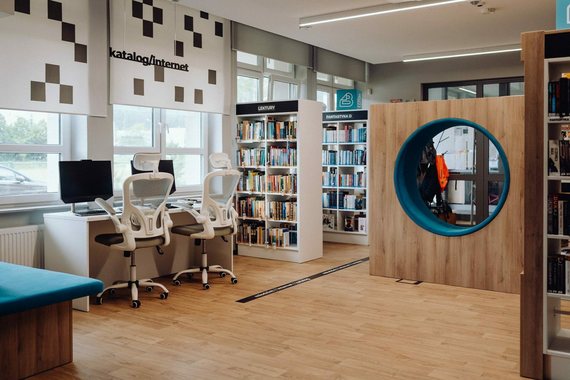 The modern library with wooden floor is equipped with white bookcases filled with books. Two ergonomic chairs and computer desks are located near large windows. A large round blue reading nook is integrated into the wooden structure, providing a cozy reading space perfect for relaxing after a photography event.  