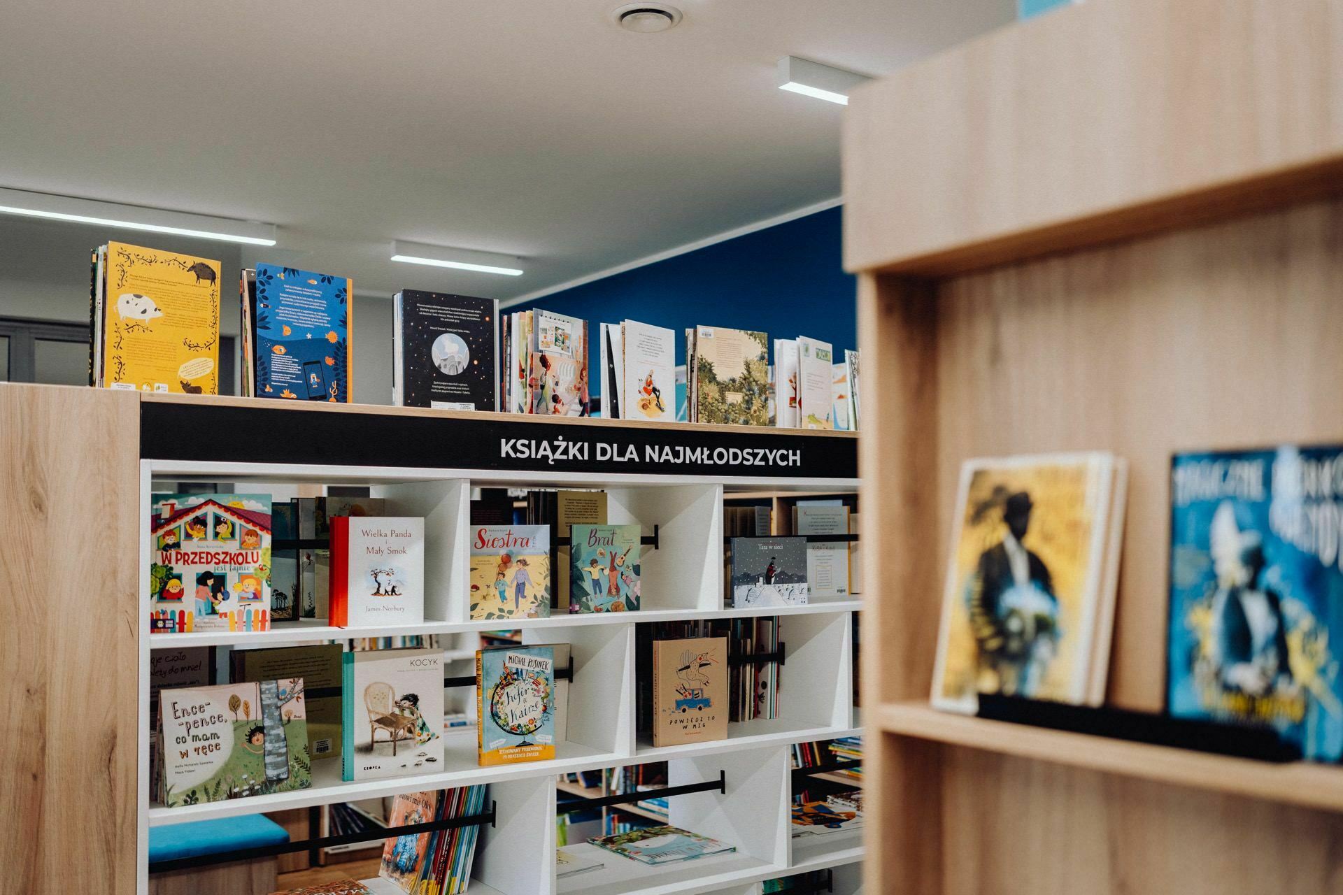 A well-lit section of a children's bookstore with colorful books displayed on white shelves. On the shelves, an upper sign in Polish reads "KSIĄŻKI DLA NAJMŁODSZYCH", meaning "Books for the youngest". A Warsaw photographer captured this charming space, which also has books on an adjacent wooden shelf.  