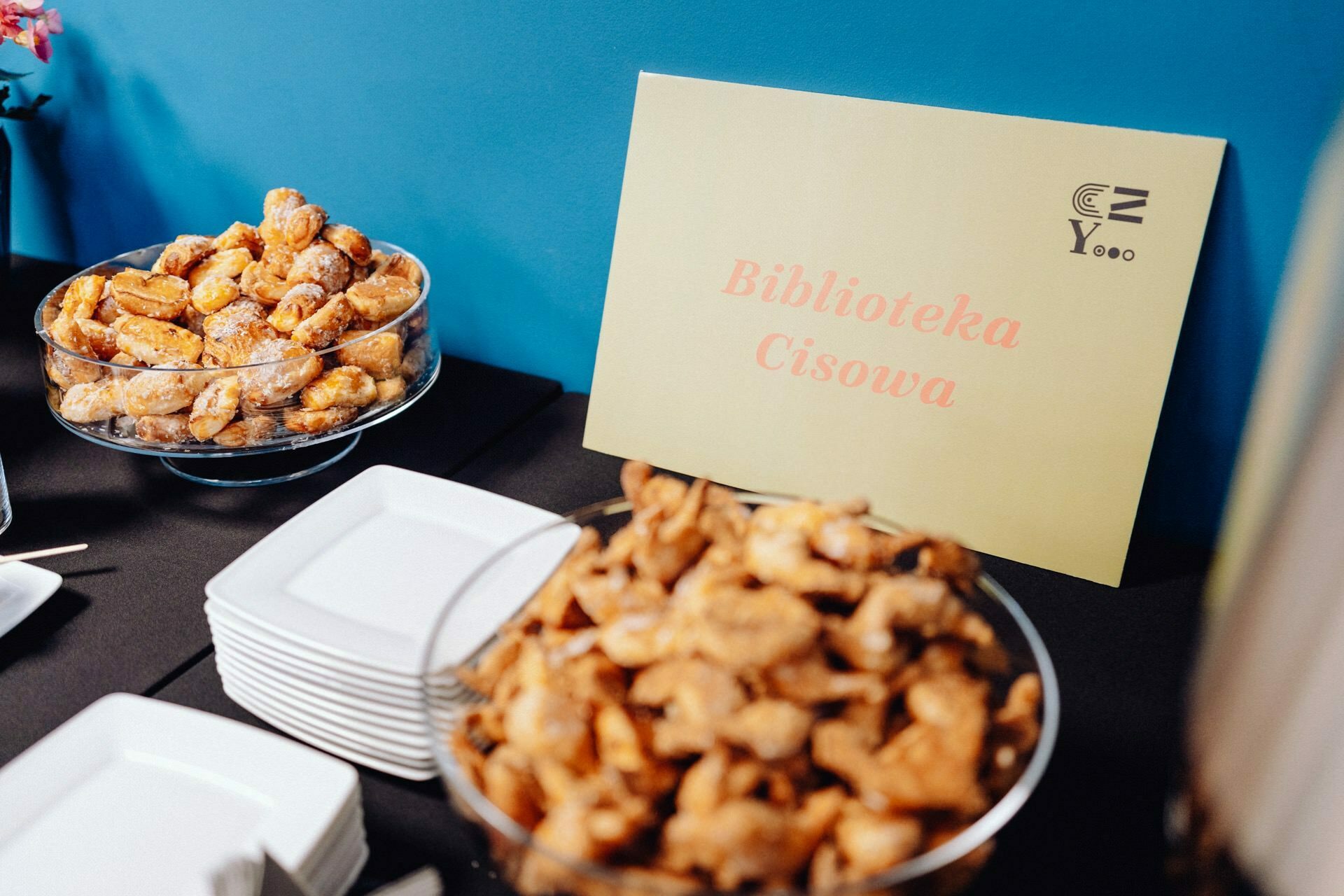 A table with trays of cakes and stacks of square white plates. In the background is a pastel-colored signboard that reads "Cisowa Library." The scenery suggests a food setting, perhaps for a party or meeting, which was perfectly captured in a photo report of the event by the talented event photographer Warsaw. Behind them is a serene blue wall.   