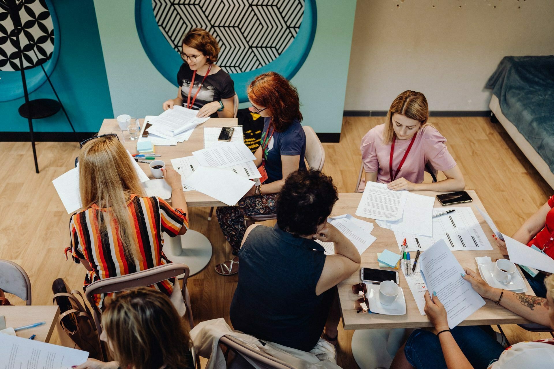 A group of people are sitting at a table having a discussion. Various documents, papers and cups of coffee lie on the table. The setting appears to be a casual, modern work space with light wooden floors, blue walls and geometric wall decoration - perfect for a photojournalism of the event by an experienced event photographer warsaw.  