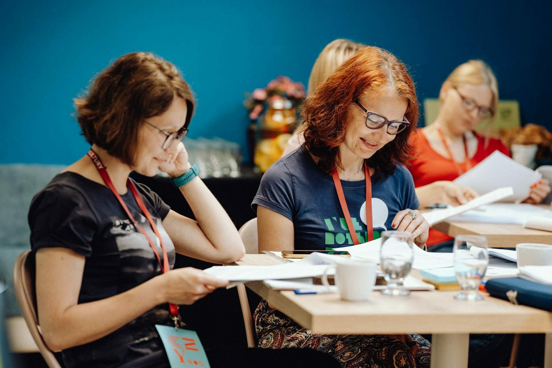 Four women sit at tables in a room with blue walls. They look focused, reading and writing on pieces of paper. One woman has red hair and glasses, another has short brown hair and glasses. Drinks and notes lie on the table. The studio atmosphere was captured by a photographer from Warsaw doing a photo report of the event.    