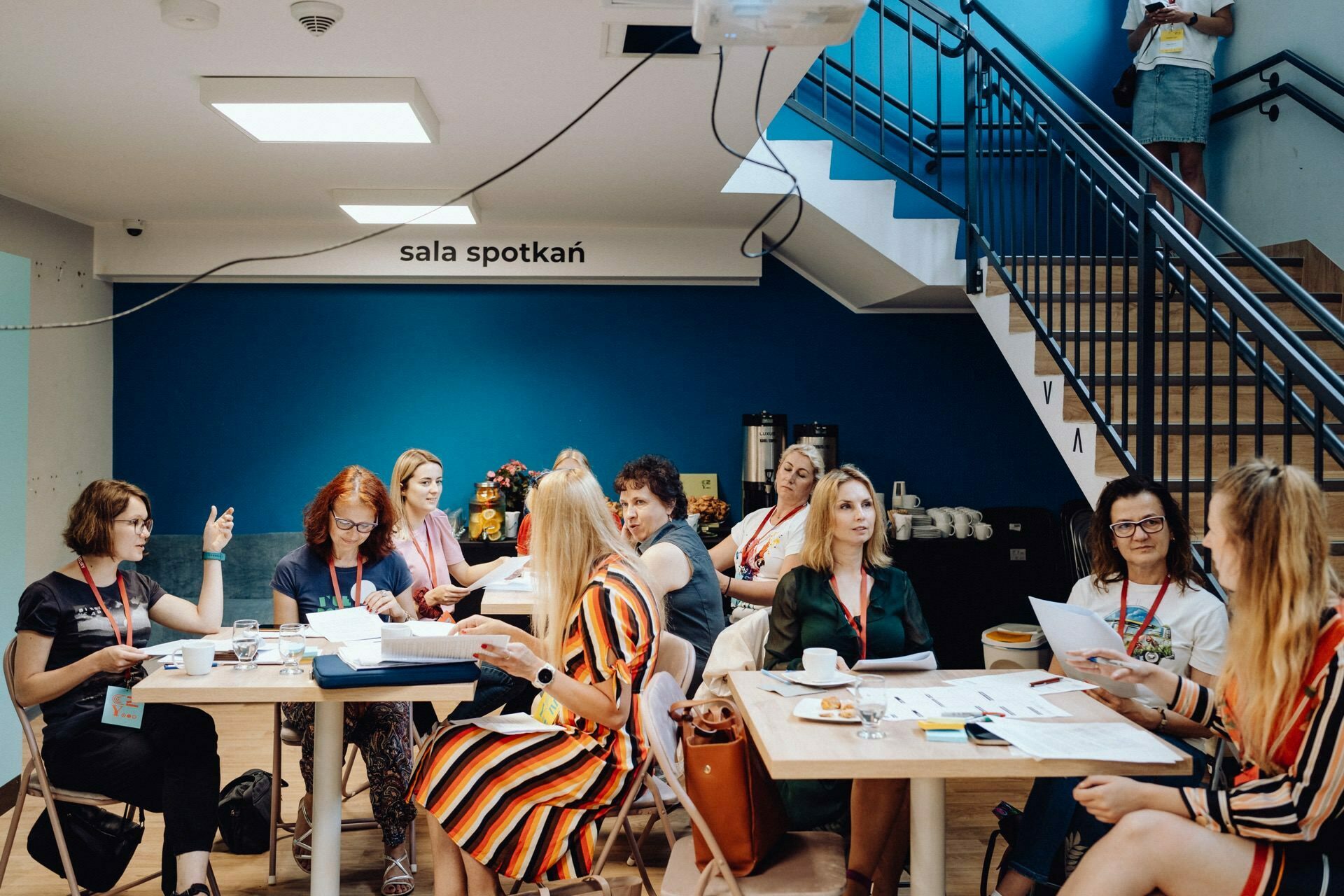 A group of women sit around tables in a brightly lit conference room with a blue wall labeled "meeting room" (meeting room). They are having a discussion, some holding papers and carrying lanyards. A staircase is visible in the background, beautifully captured by a photographer from Warsaw.  