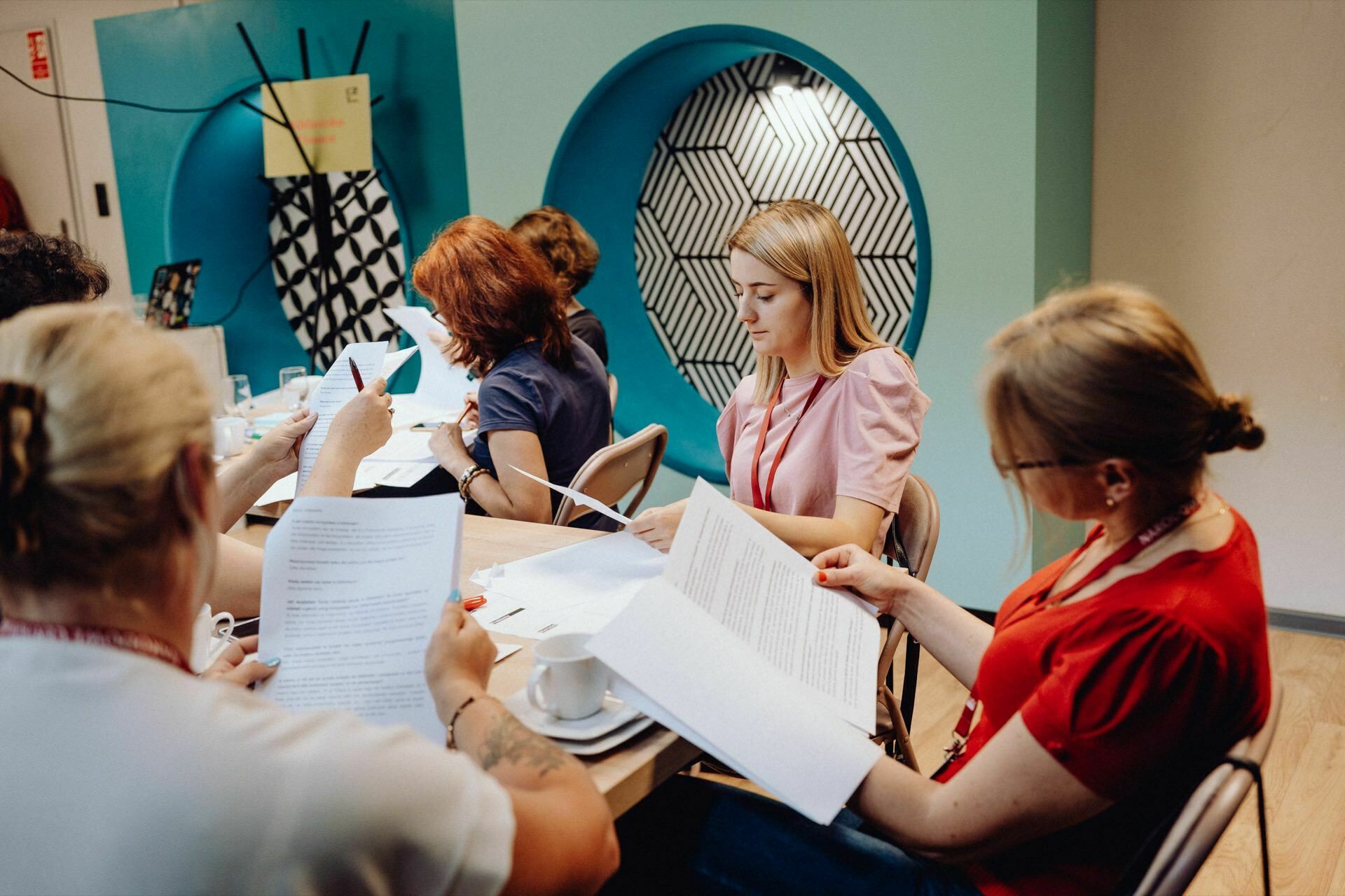 A group of people sitting at a table and reading documents. The place looks like an ordinary office or conference room with turquoise round wall decorations and patterned designs. The event photographer Warsaw captured the atmosphere of concentration as everyone peruses their papers, drinks on the table.  