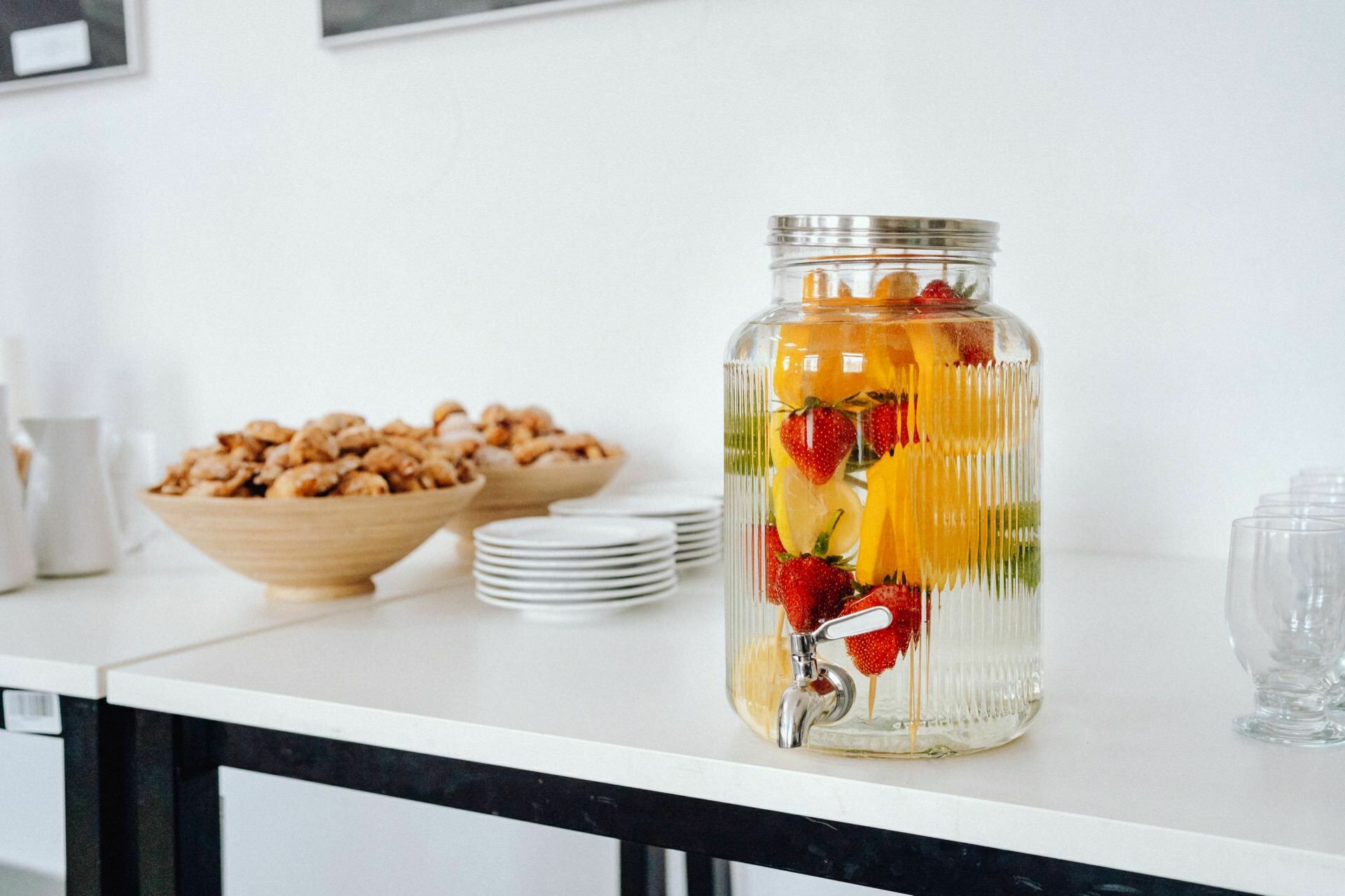 A beverage dispenser filled with water topped with fruit, including oranges and strawberries, stands on a white countertop. Stacked plates, clear glasses and two wooden bowls filled with cakes stand nearby. In the background of the photo event is a white wall decorated with framed photos.  