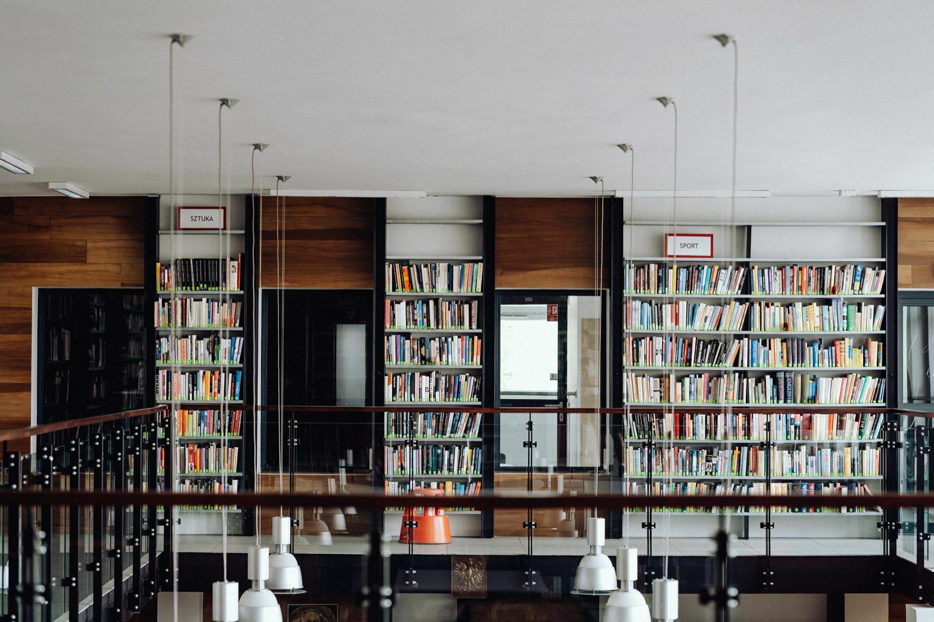 The spacious interior of the library features tall shelves filled with books, neatly organized into sections. Modern white pendant lamps hang from the ceiling. Above the shelves are signs reading "ART" and "SPORT." The well-known event photographer warszawa perfectly captures the serene and orderly atmosphere.   