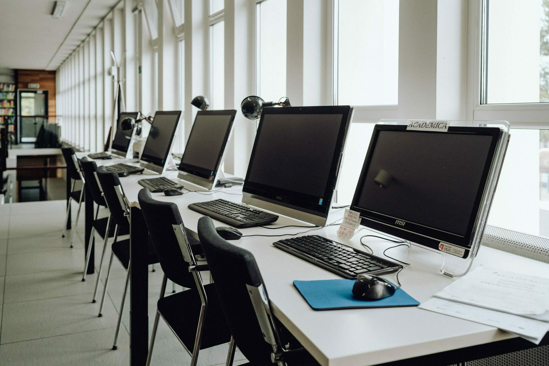 A row of desktop computers with keyboards and mice stand on a long white desk in a well-lit room, probably in a library or computer lab. A black chair sits at each workstation, and windows run along the wall, letting natural light into the room - the perfect backdrop for any Warsaw photographer recording an event. 