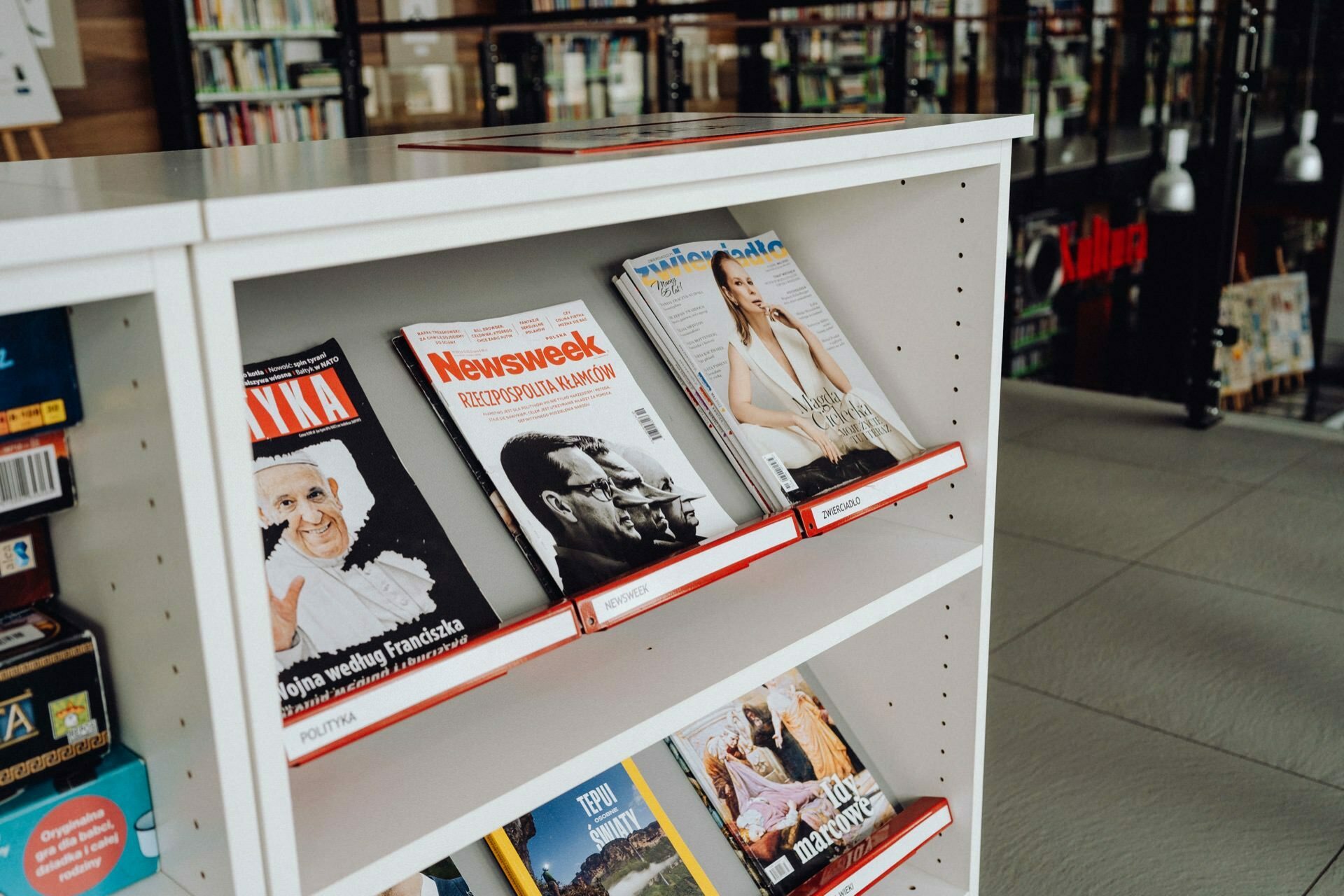 A white bookcase in a bookstore or library displays various magazines. The top shelf holds "Newsweek" and other popular magazines. In the background, books are stacked on the shelves, and hanging lamps add a warm glow, creating an atmosphere perfect for a photo event.  