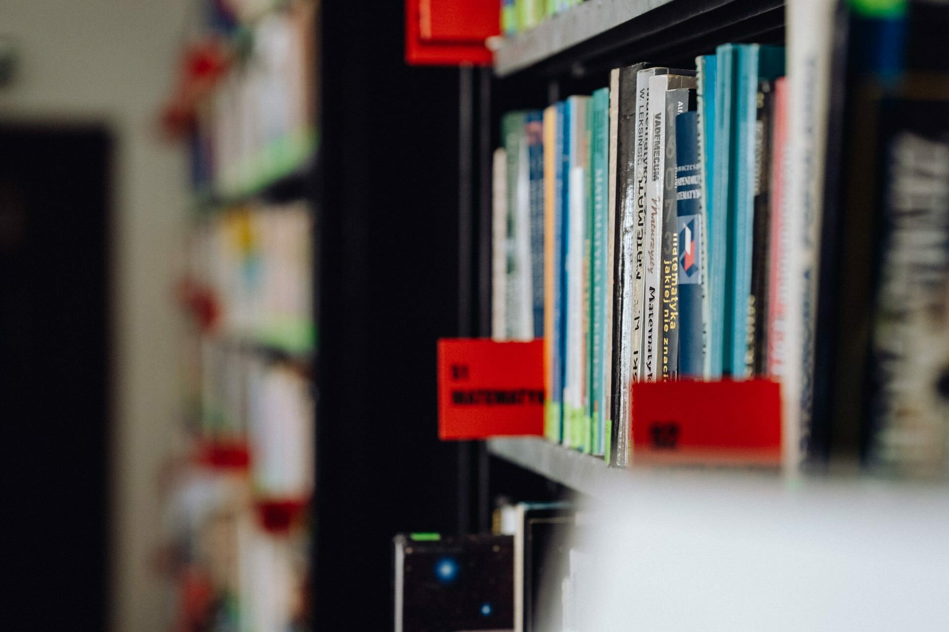 A close-up view of library bookshelves captured by a photographer from Warsaw. The foreground shows a row of colorful books with red labels indicating their categories, while the background is deliberately blurred, focusing attention on the neatly organized books and their vibrant covers. 