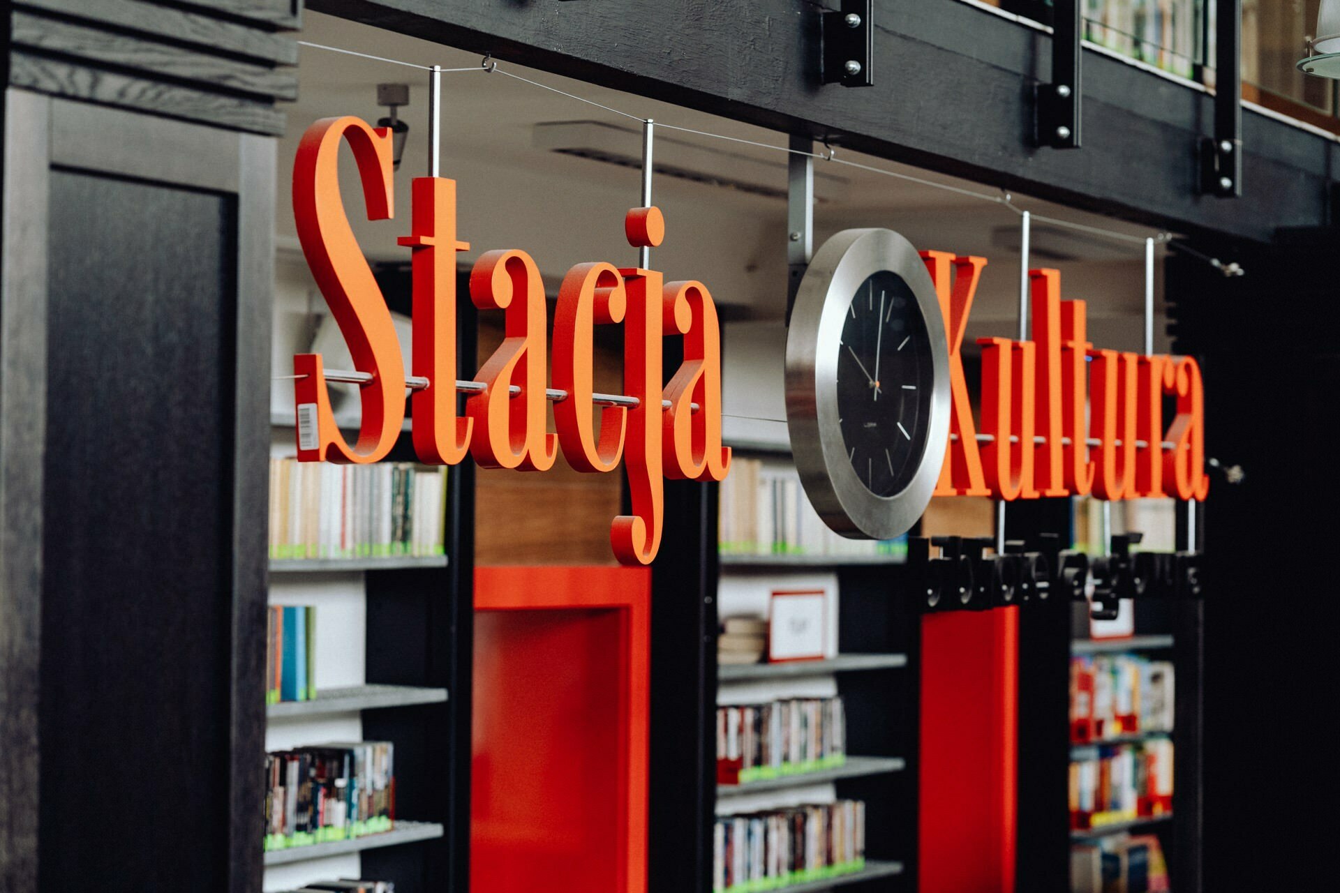 The interior of the library with a large sign with the words "Culture Station" in red letters hanging from the ceiling. Below the sign are shelves filled with books. Between the words is a modern, circular black and white wall clock - perfect for photo coverage of the event by any event photographer Warsaw.  